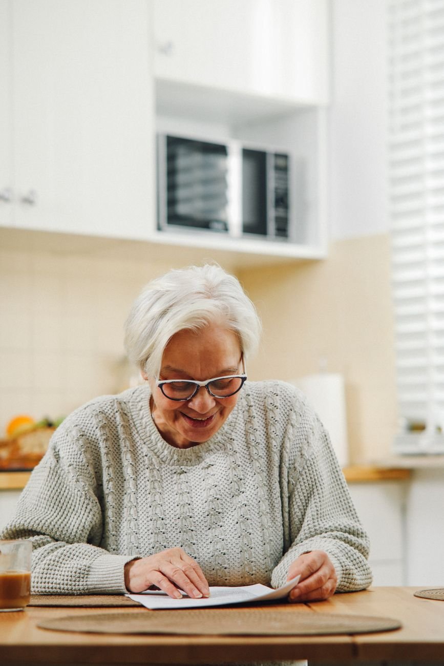 A smiling elderly woman with white hair reading a paper at her kitchen table, enjoying a serene morning.